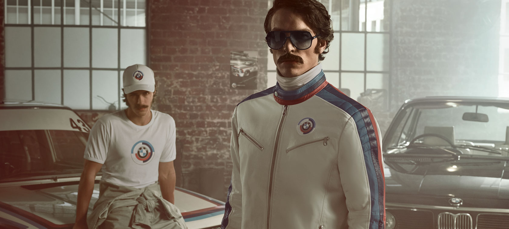 BMW Lifestyle. Man met iconisch BMW Motorsport Heritage leren jack – op de achtergrond een tweede man in kleding van BMW Heritage.